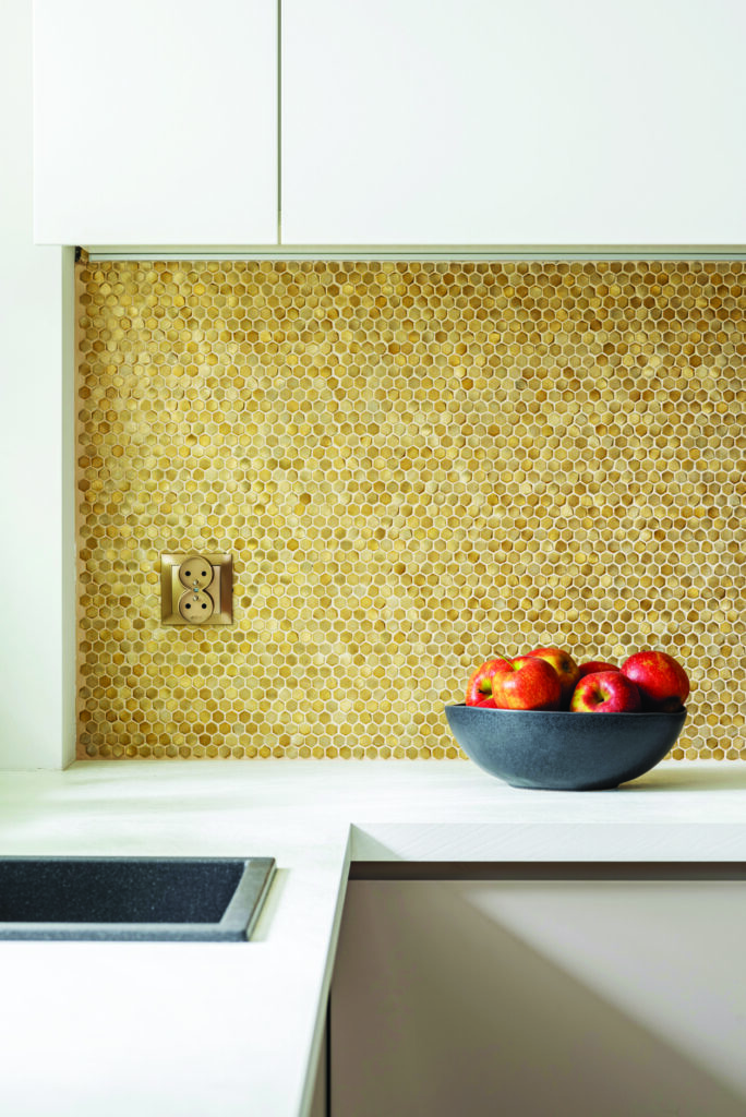 Das DUNIN Allumi Gold Hexagon Wandmosaik dient als dekorative Oberfläche für eine Küchenwand. Die goldenen Fliesen harmonieren perfekt mit den hellen Schränken und der modernen Arbeitsplatte.